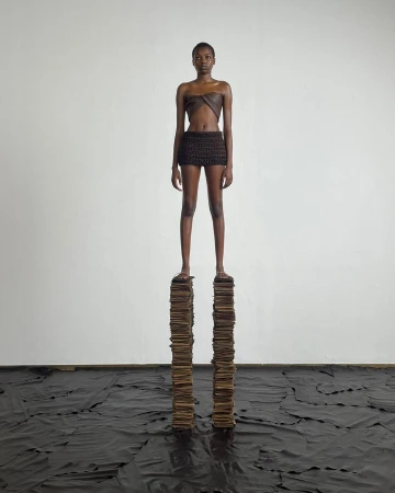 A slender woman in a brown two-piece outfit confidently stands on a stack of books indoors against a white wall, with a dark material on the floor.
