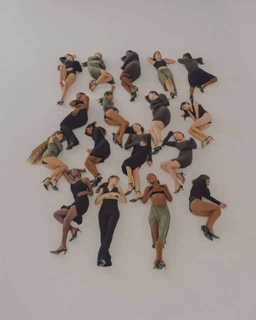 High-angle shot of a diverse group of women lying in a circle on a plain white surface, posing in various outfits and elegant heels.