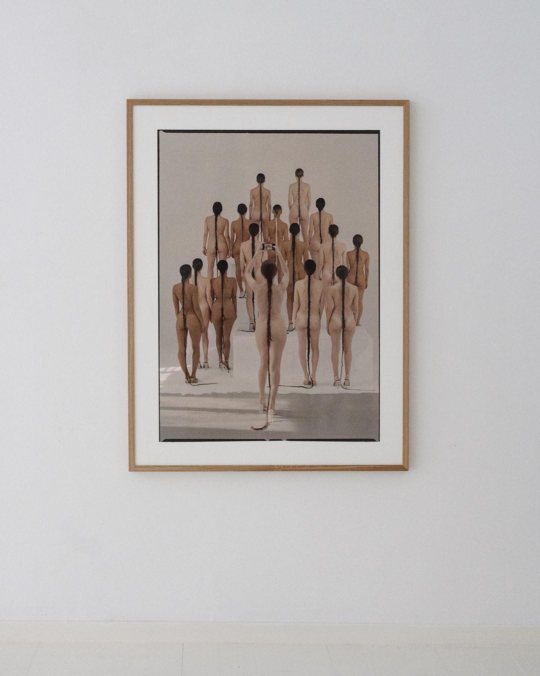 Artistic photograph of a framed print depicting a group of nude figures, seen from behind, with hair braided down their backs.