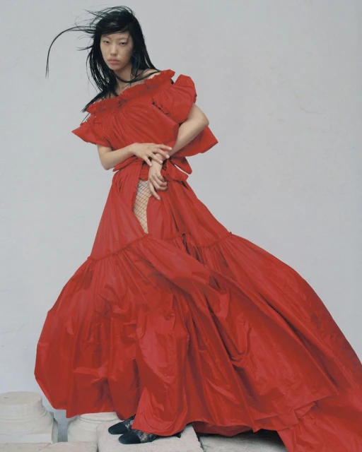 A woman posing in a dramatic red gown with ruffles and tulle, set against a neutral backdrop, captured in a fashion photography style.