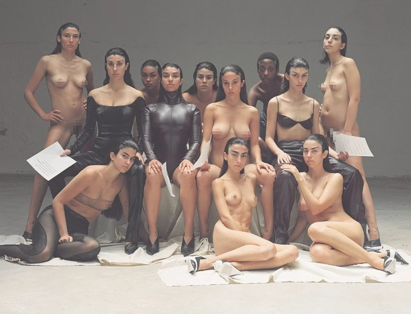 Group of nude and partially clothed women in black outfits pose in a studio setting, holding sheets of paper in a staged setting.