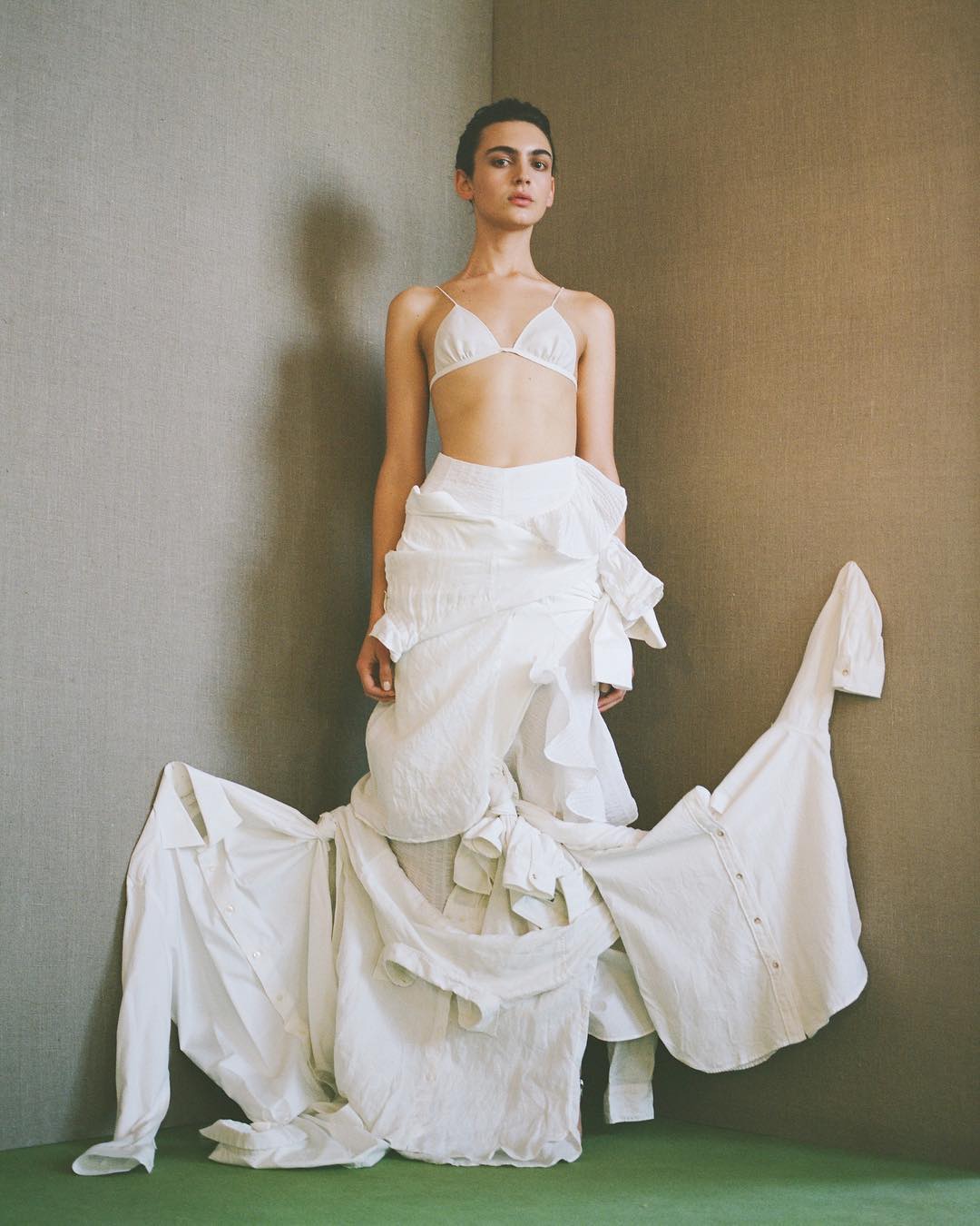 Fashion shot of a young adult woman in a white bra top and white shirts assembled as a skirt, posing indoors in a corner setting.