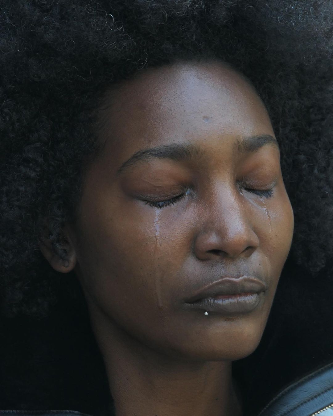 Close-up portrait of a crying African woman with closed eyes, tears on her face, and afro hairstyle in a dark outdoor setting.