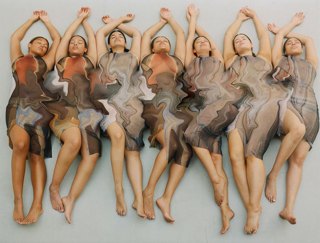 A group of six adult women lying side-by-side in a studio, wearing patterned dresses, eyes closed, and arms raised in an artistic pose.