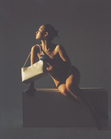 Woman in profile seated on a block holding a white handbag with blue gloves under dramatic studio lighting by Carlota Guerrero.