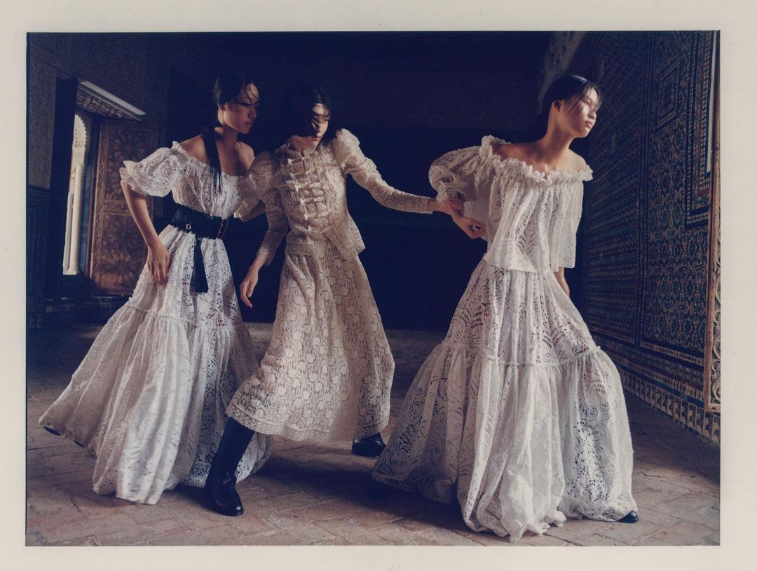 Three women pose indoors wearing lace dresses, shot at eye level, with dark background and a floor made of brown tiles.