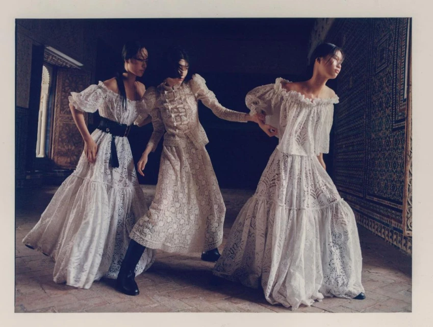 Three women pose indoors wearing lace dresses, shot at eye level, with dark background and a floor made of brown tiles.