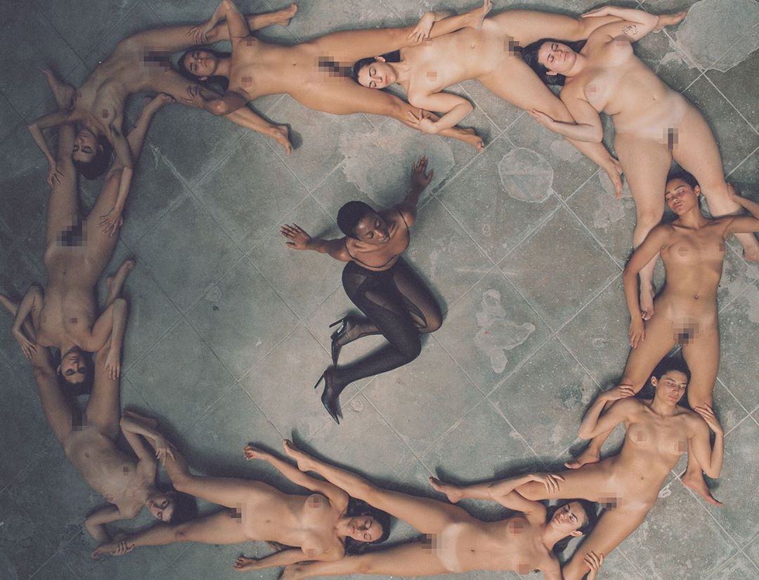Overhead shot of a woman in dark tights surrounded by nude women forming a circle on a tiled floor, casting a shadow effect.