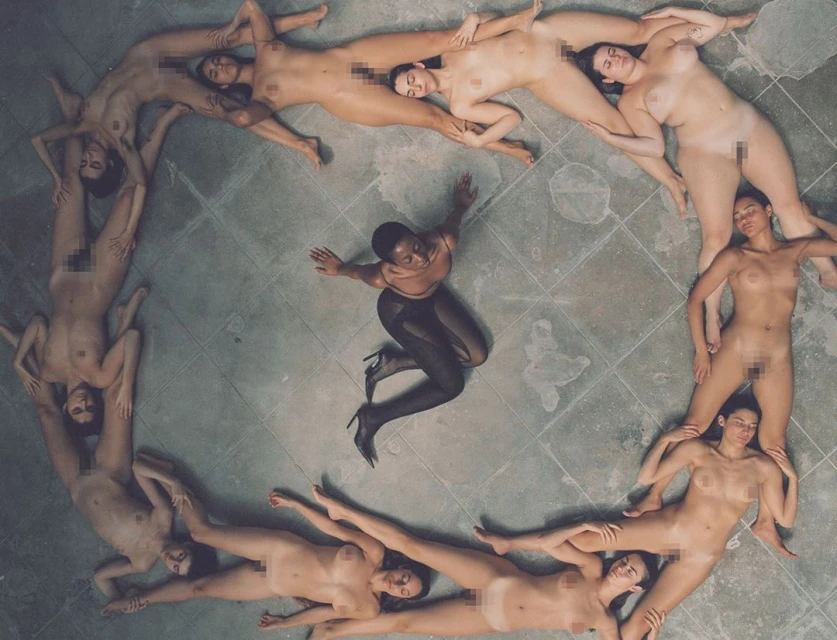 Overhead shot of a woman in dark tights surrounded by nude women forming a circle on a tiled floor, casting a shadow effect.