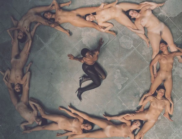 Overhead shot of a woman in dark tights surrounded by nude women forming a circle on a tiled floor, casting a shadow effect.
