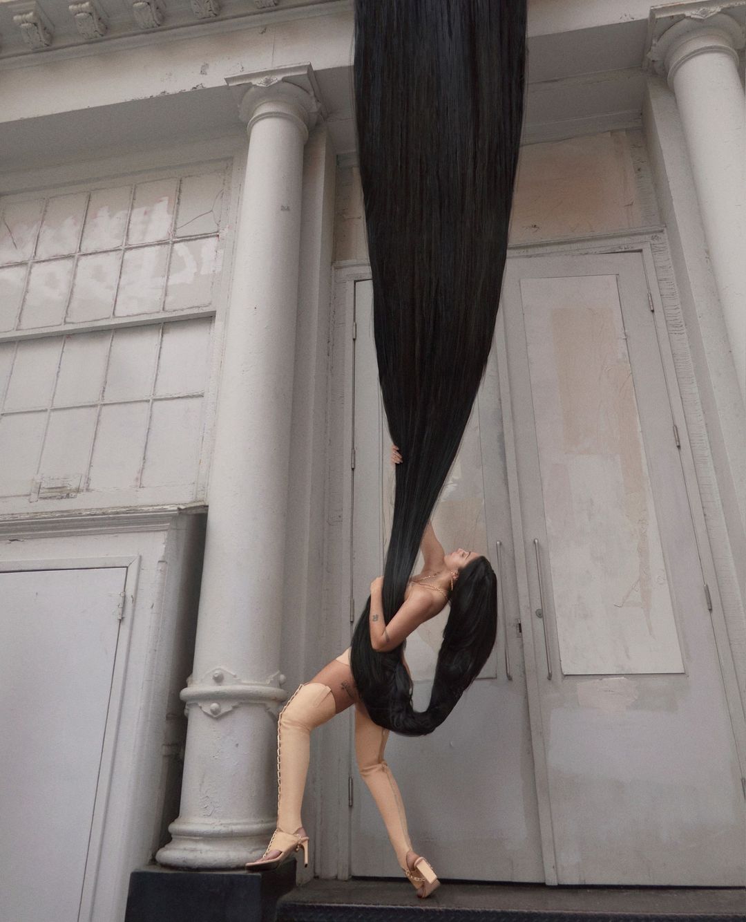 Woman with extremely long black hair stretching upward into the sky against a classical building exterior with columns