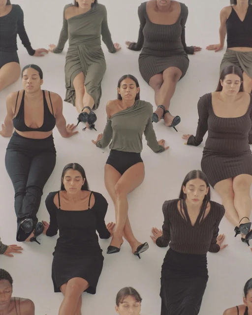 Group of diverse women in black and olive-green dresses and outfits sit in a meditative pose against a neutral white backdrop.
