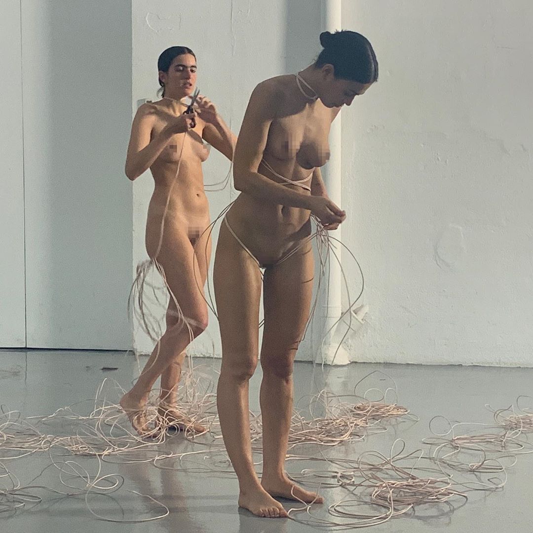 Two nude women are standing indoors surrounded by pink wires. One woman is cutting a wire with scissors, and the other is adjusting wires around her.