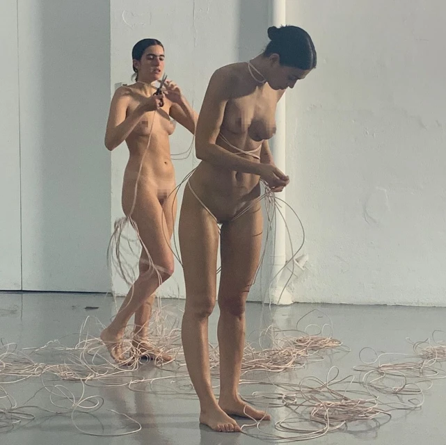 Two nude women are standing indoors surrounded by pink wires. One woman is cutting a wire with scissors, and the other is adjusting wires around her.
