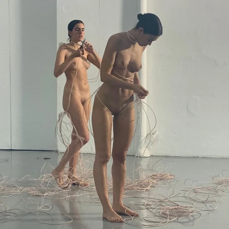Two nude women are standing indoors surrounded by pink wires. One woman is cutting a wire with scissors, and the other is adjusting wires around her.