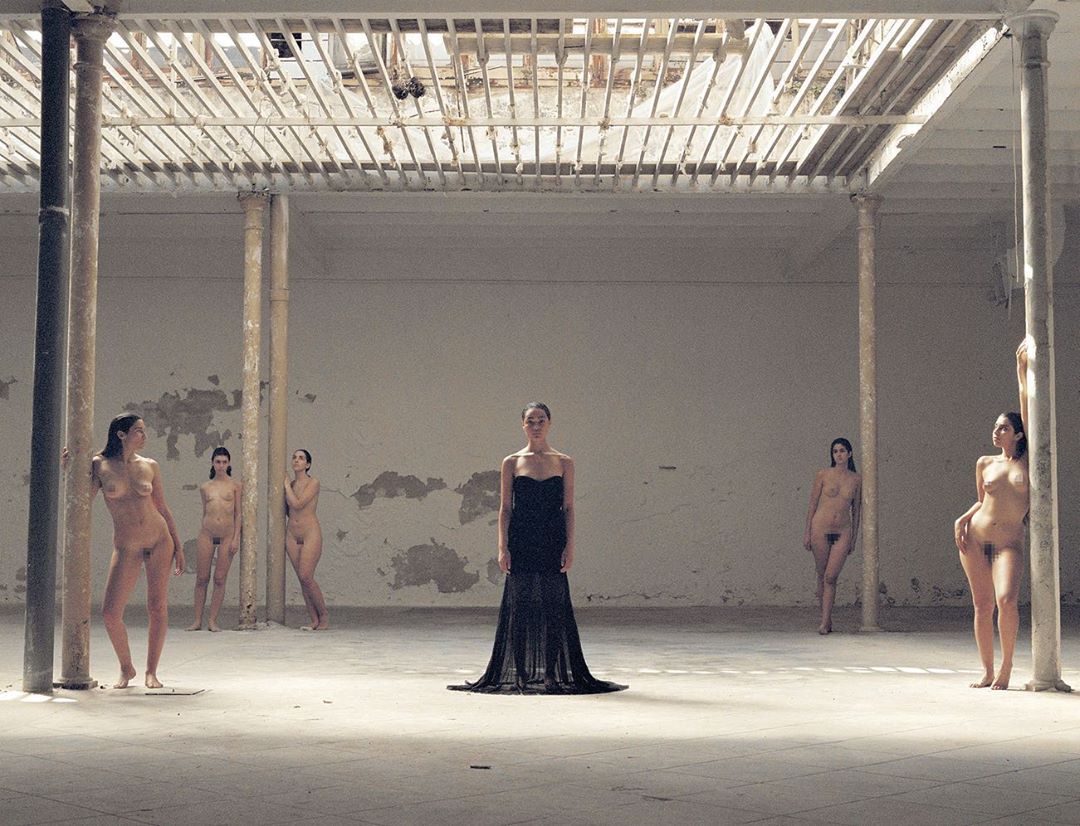 A woman in a black gown stands in a spacious hall, surrounded by partially nude women, under a high ceiling with white support beams.