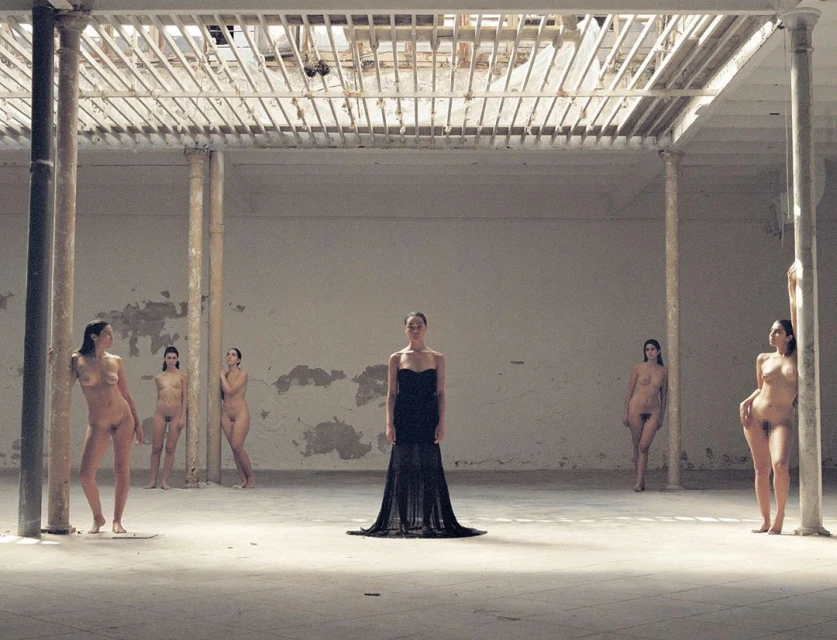 A woman in a black gown stands in a spacious hall, surrounded by partially nude women, under a high ceiling with white support beams.