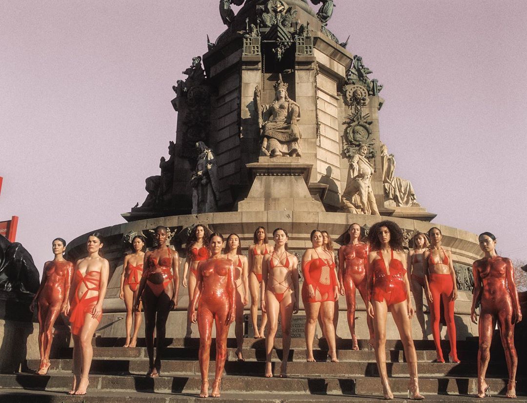 A group of diverse women in red body paint stand on steps in front of a monument in an outdoor urban environment under a bright sky.