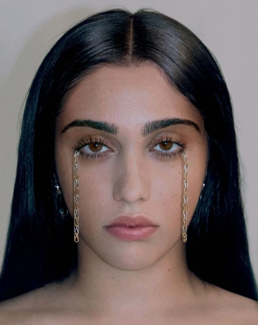 Close-up portrait of a woman with dark hair and brown eyes, featuring gold chains styled as tears under her eyes, against a neutral backdrop.