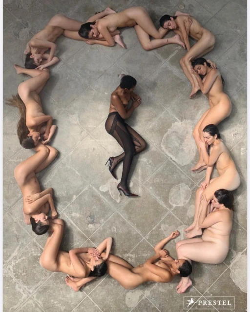 Overhead shot of eleven nude women forming a circle around a woman in black tights and heels, on a textured gray concrete floor.