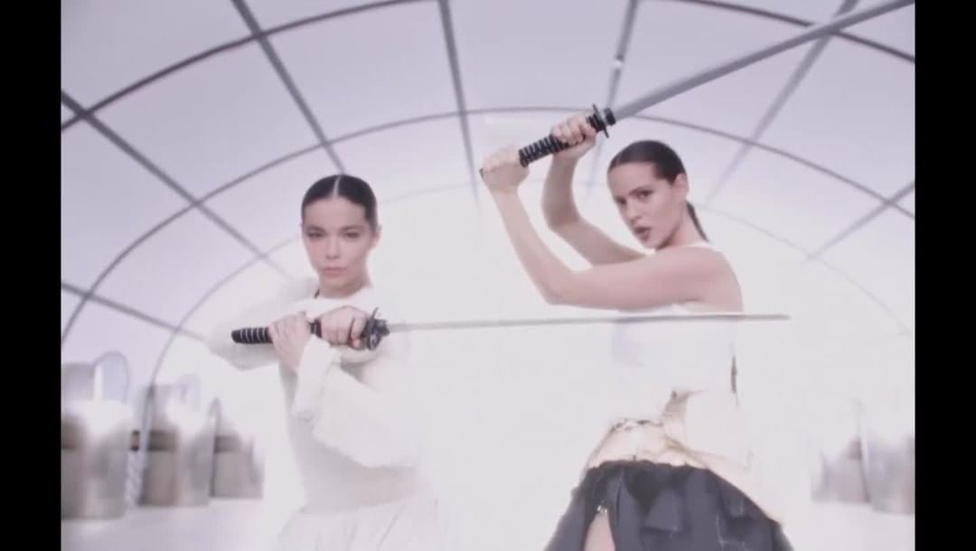 Two adult women holding swords in a brightly lit photo studio with white geometric background; fashion, beauty and lifestyle imagery.
