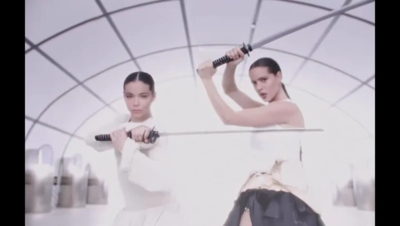 Two adult women holding swords in a brightly lit photo studio with white geometric background; fashion, beauty and lifestyle imagery.