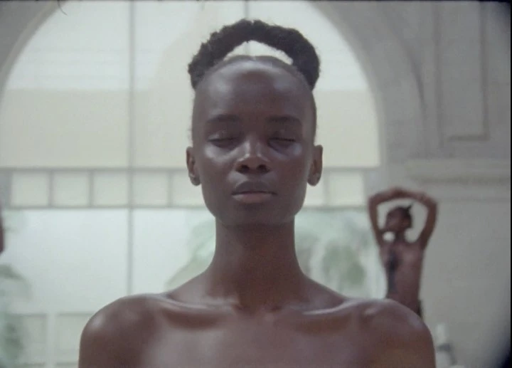 Close-up of a Black woman with hair styled in a bun and her eyes closed, along with blurry figures and a white-arched background.