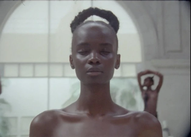 Close-up of a Black woman with hair styled in a bun and her eyes closed, along with blurry figures and a white-arched background.