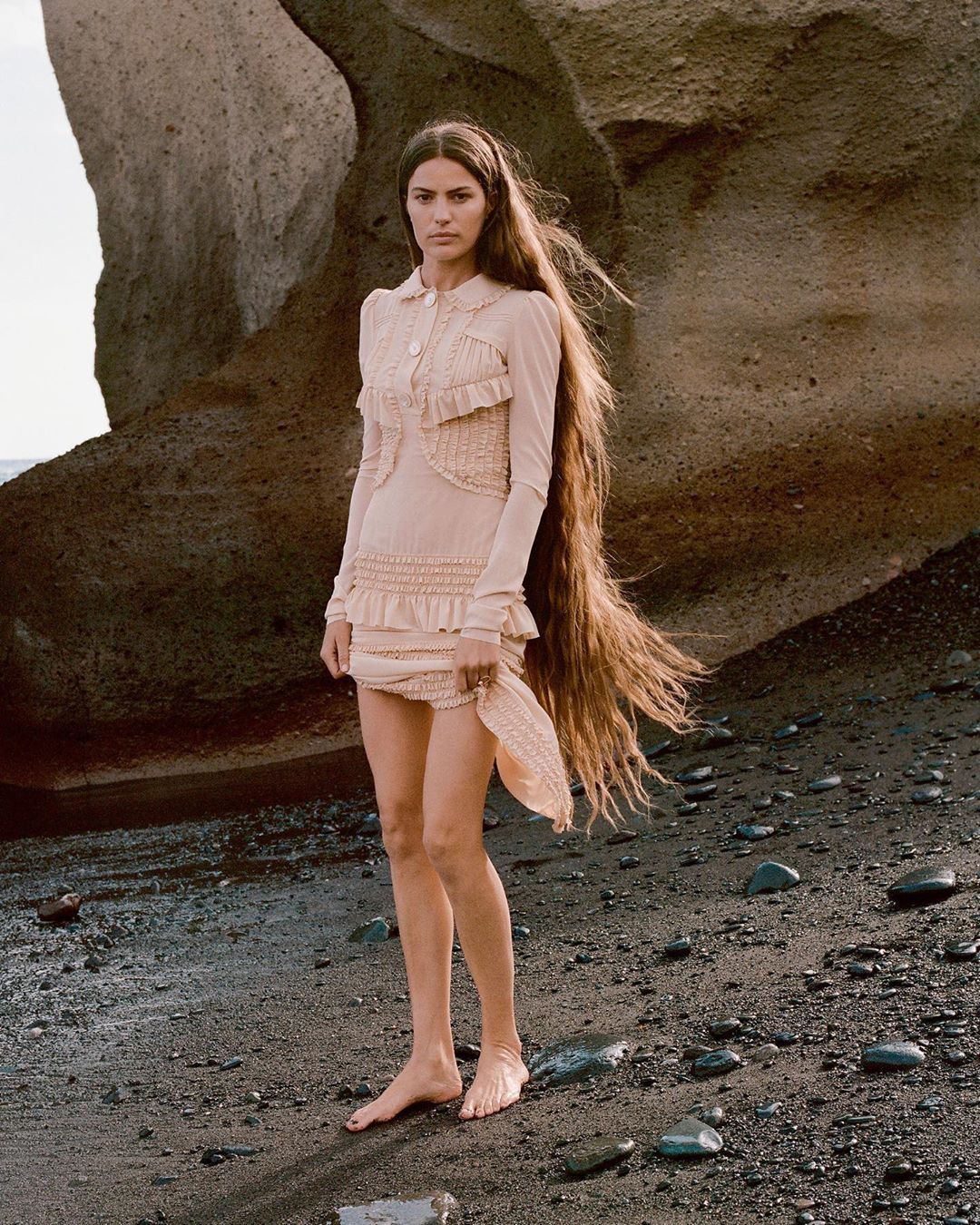 An adult female model stands barefoot on a dark sand beach in a light tan dress with long brown hair, posing in front of a large rock.