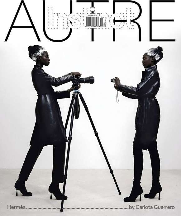 Two dark-skinned women in black outfits photograph each other in a white studio, one using a tripod-mounted camera.