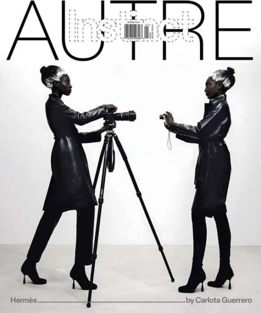 Two dark-skinned women in black outfits photograph each other in a white studio, one using a tripod-mounted camera.