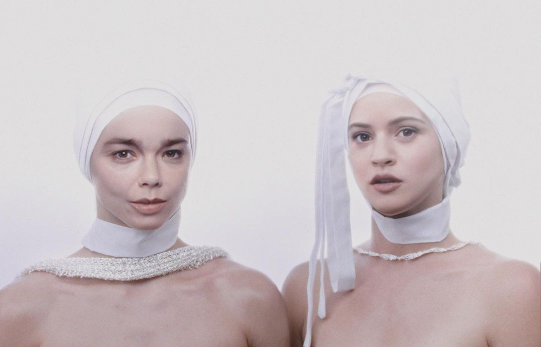 Two adult women face the camera in a portrait style, wearing white fabric head coverings and simple neckwear against a white backdrop.