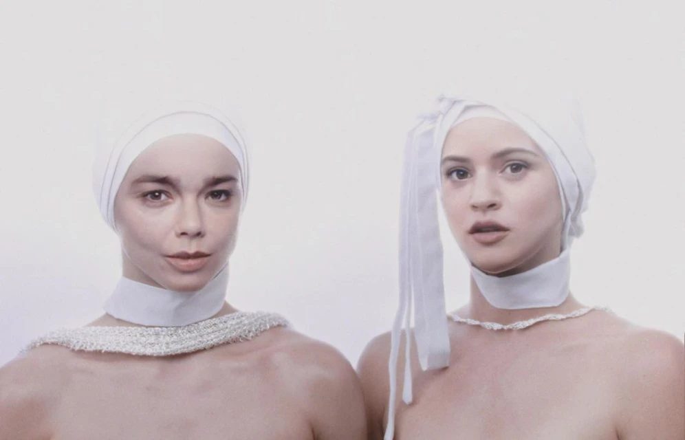 Two adult women face the camera in a portrait style, wearing white fabric head coverings and simple neckwear against a white backdrop.