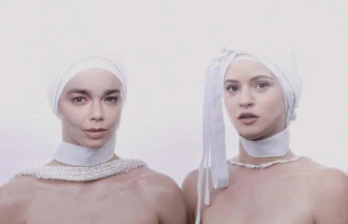 Two adult women face the camera in a portrait style, wearing white fabric head coverings and simple neckwear against a white backdrop.