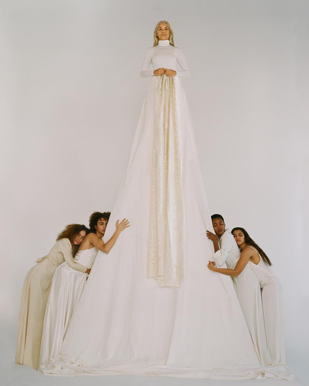 A woman in a long white dress stands tall with five people supporting the dress around her in a light, airy studio setting.