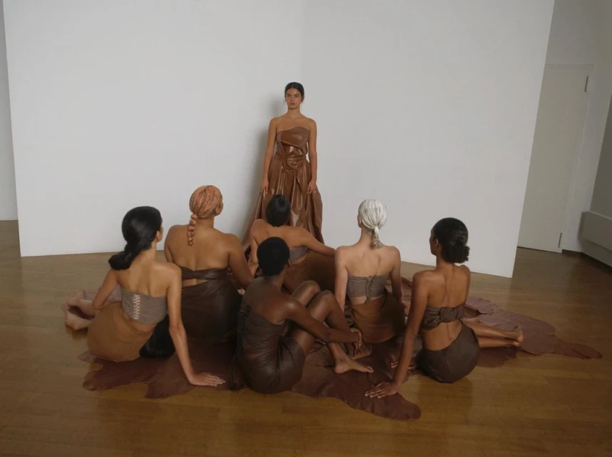 Group of seven women in draped brown fabric in photo studio. One stands; others sit in a semi-circle. Minimalist fashion and art.