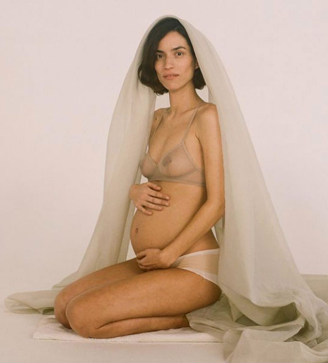 Pregnant woman kneeling, draped in sheer fabric, in a studio setting. Soft lighting highlights her form and the fabric's delicate texture.
