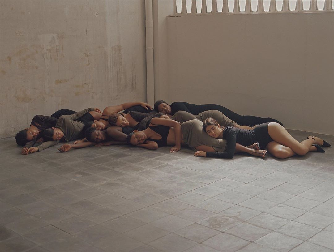 Large group of multi-ethnic adults lie close together on the floor of an empty studio with light grey walls and a tiled floor.