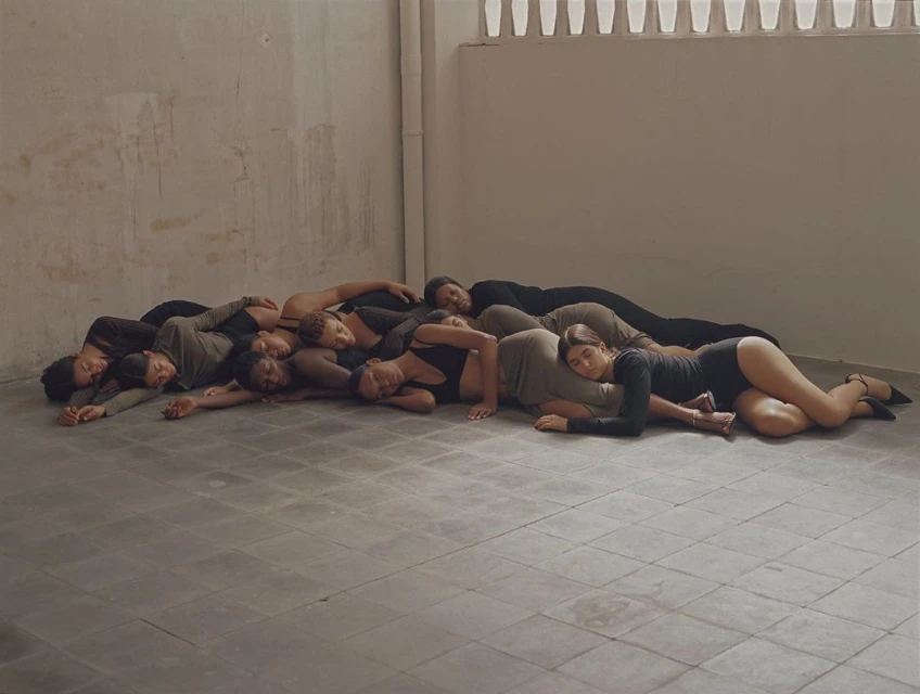 Large group of multi-ethnic adults lie close together on the floor of an empty studio with light grey walls and a tiled floor.