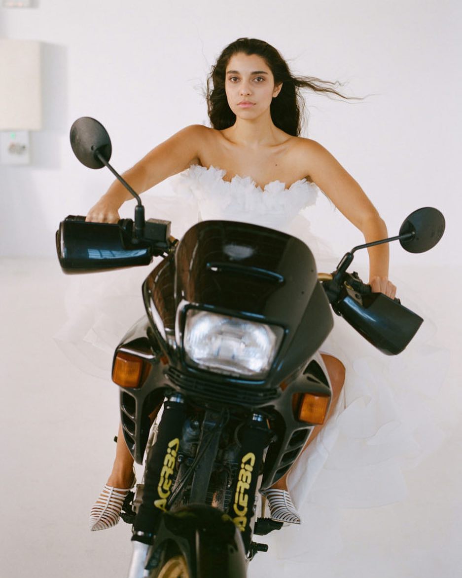 Woman in tulle dress sits astride a black motorcycle in a photo studio setting, illuminated by soft, natural light. 