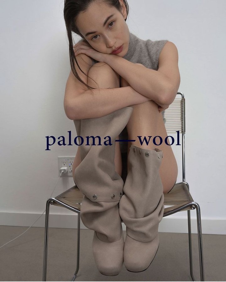 An adult female sits on a chair wearing boots in a studio with a neutral wall, photographed in a medium shot with soft lighting.