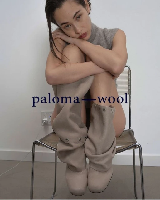 An adult female sits on a chair wearing boots in a studio with a neutral wall, photographed in a medium shot with soft lighting.