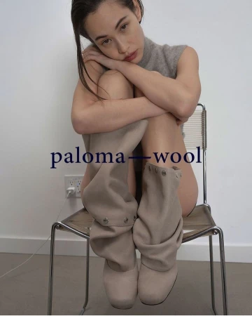 An adult female sits on a chair wearing boots in a studio with a neutral wall, photographed in a medium shot with soft lighting.
