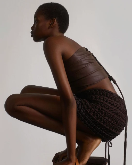 A dark-skinned woman is squatting indoors, wearing a brown leather strapless top and a brown crochet skirt against a plain background.