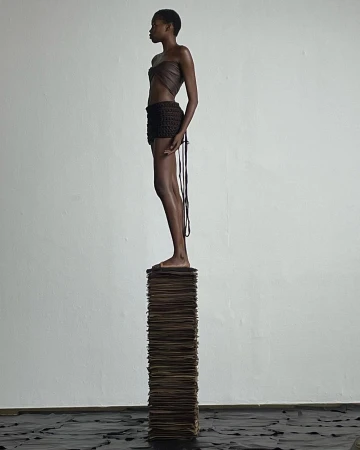 Black woman standing in profile atop a high stack of papers against a plain wall photographed by Carlota Guerrero.