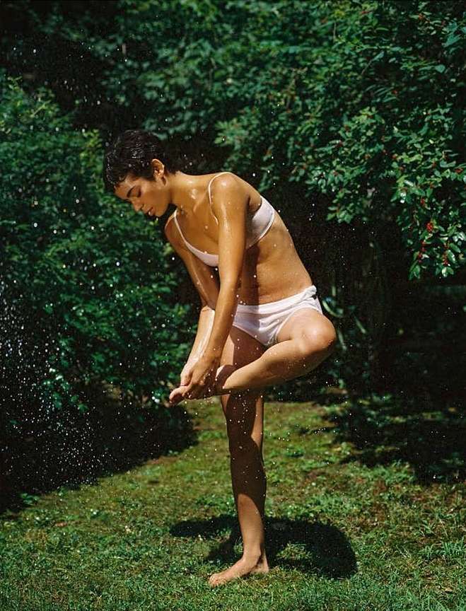 Adult woman stands in a green lawn wearing a light-pink swimsuit, water falling around her, with trees in the background on a sunny day.