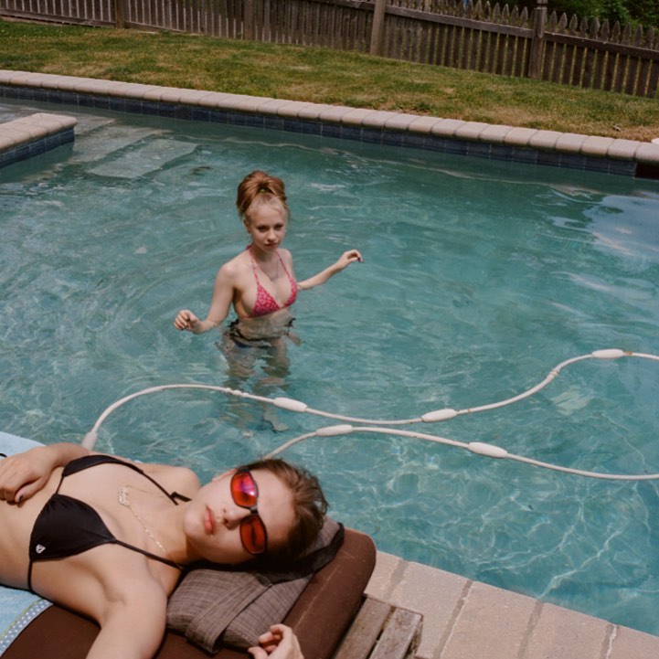 Two women in swimsuits relax at a backyard swimming pool; one woman stands in the water, the other lies on a lounge chair.