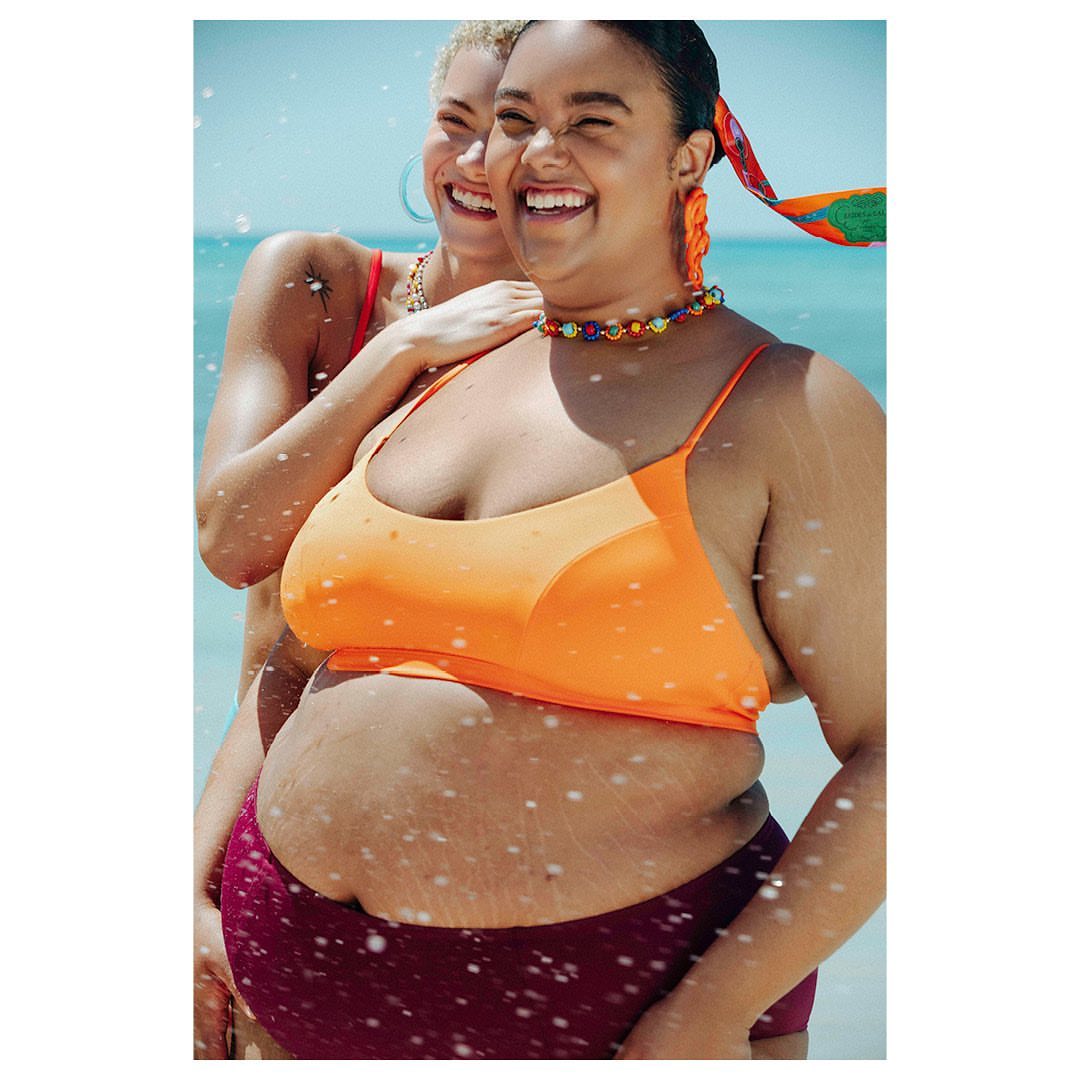Two plus-size women laugh together on a sunny beach, showcasing vibrant orange swimwear and beaded jewelry against a turquoise water backdrop.