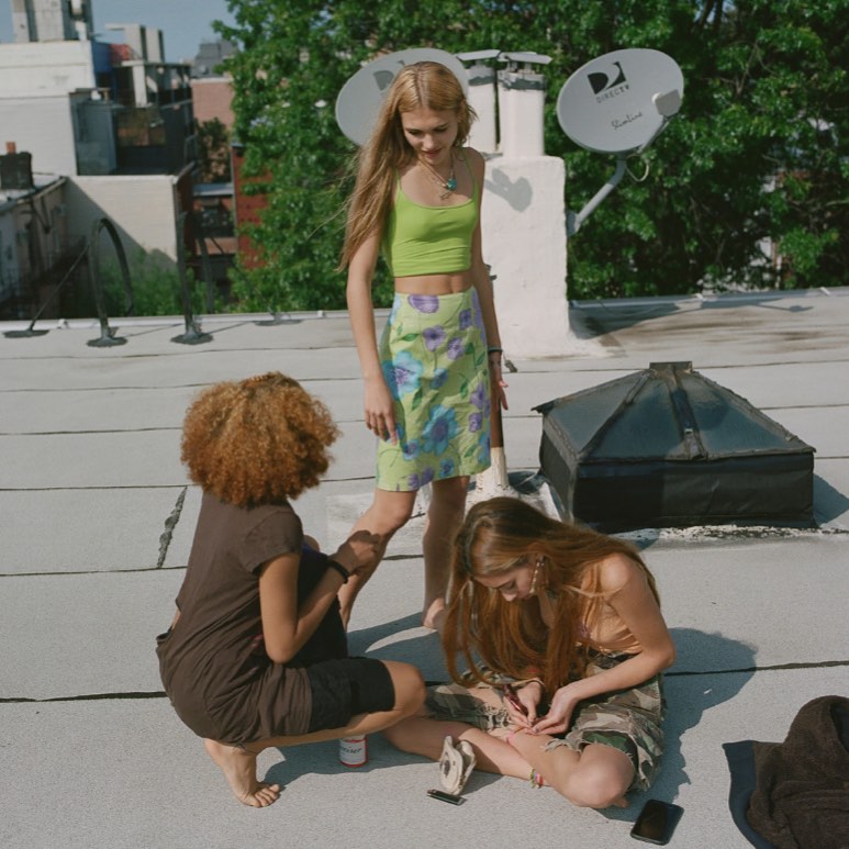 Three young women gather on a rooftop with satellite dishes and greenery in the background, two are seated while one stands.