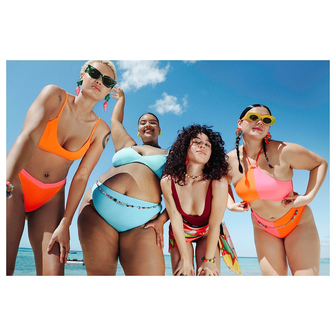 Four plus-size women in colorful swimsuits pose on a sunny beach with a blue sky, showcasing a body-positive fashion aesthetic.
